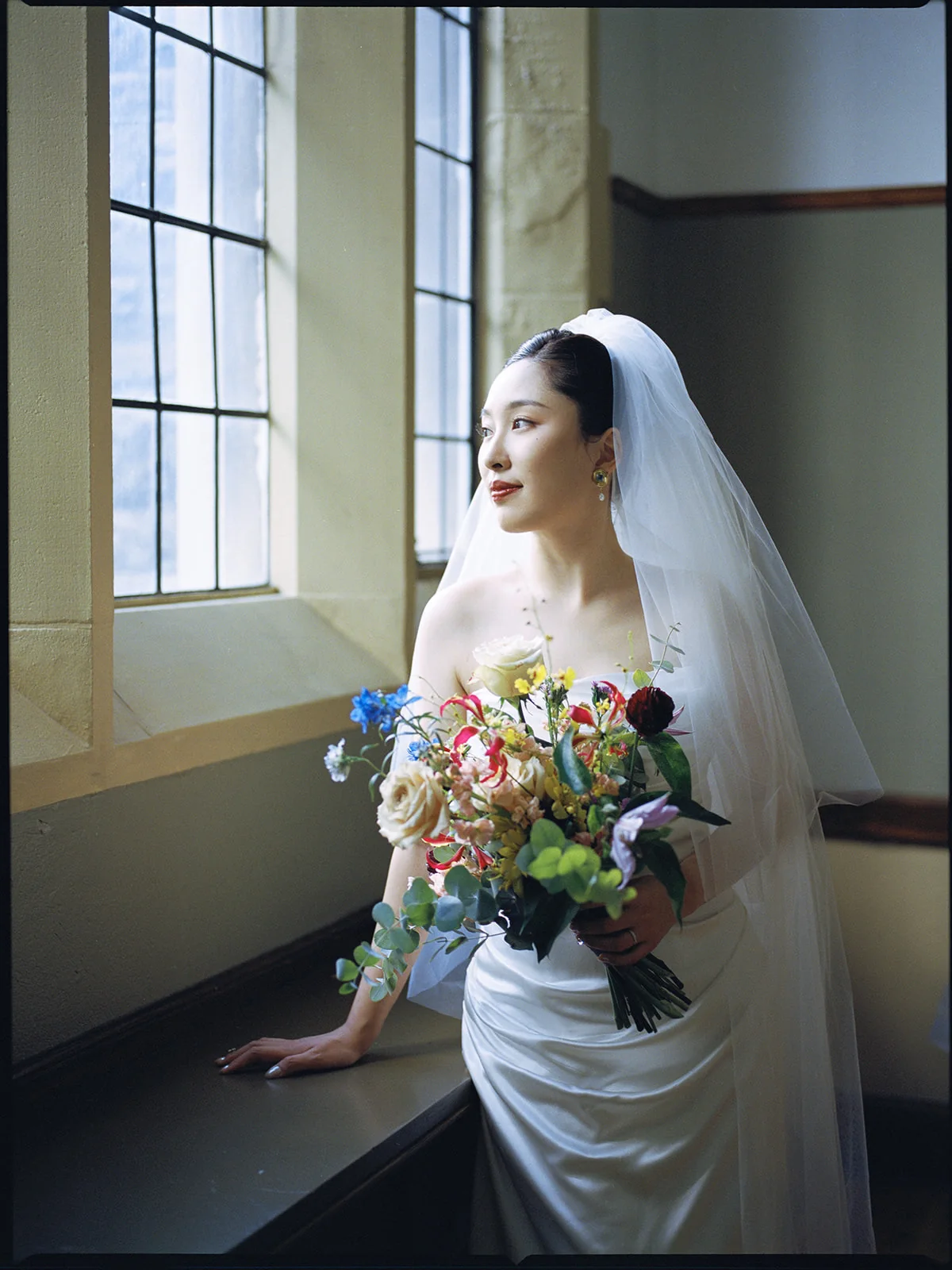Bride with a colourful bouquet beside tall windows